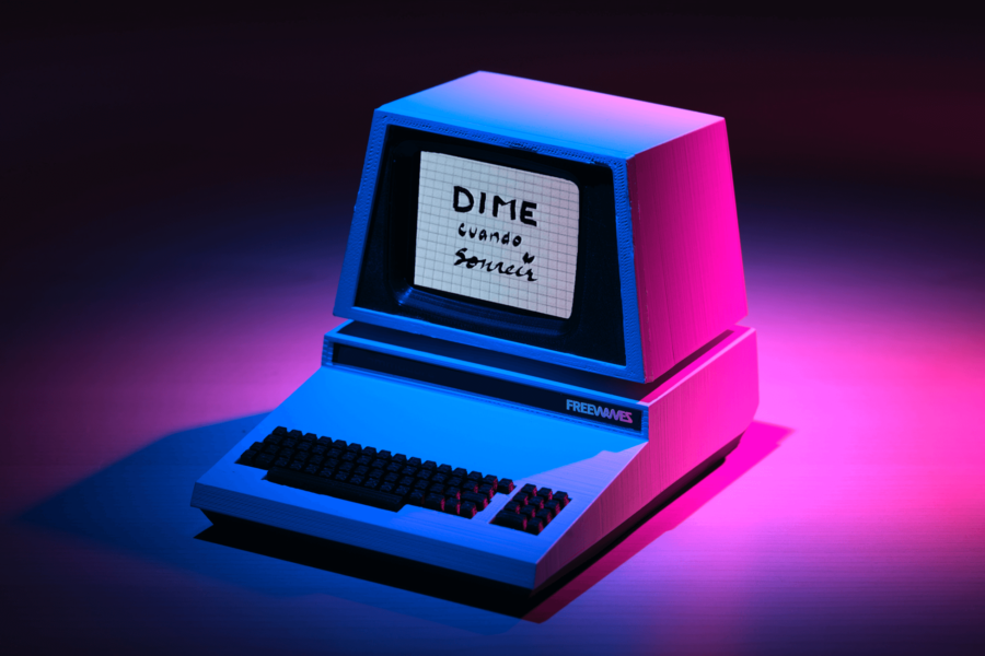 Image: Retro computer with FREEWAVES logo. The boxy monitor displays a still from an archive video titled 'DIME cuando sonreir' ('TELL me when to smile') handwritten on grid paper. The computer is on a clean surface in a dark room, illuminated by dramatic pink and blue spotlights.