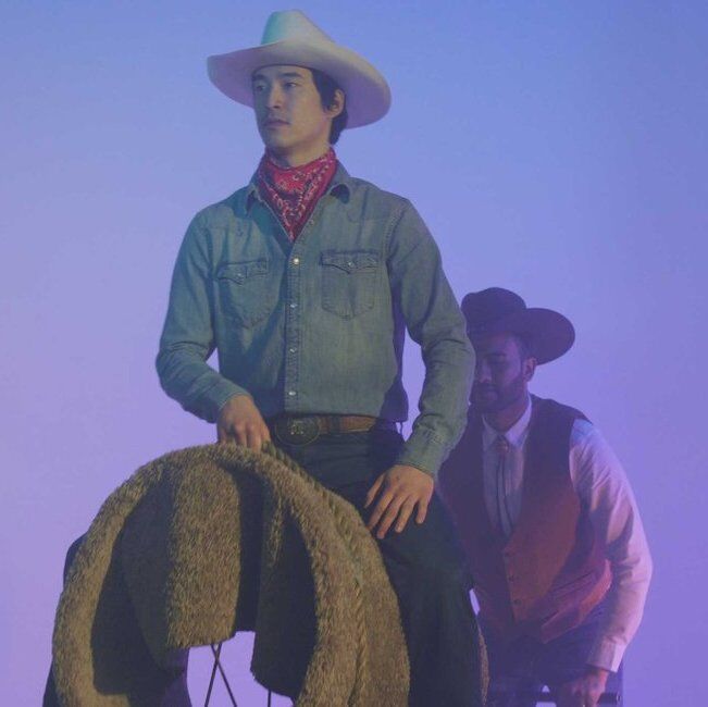 Video still of Kenneth Tam wearing cowboy attire with mounted on an invisible horse with another cowboy standing behind him. A lush lavender gradient is their backdrop.