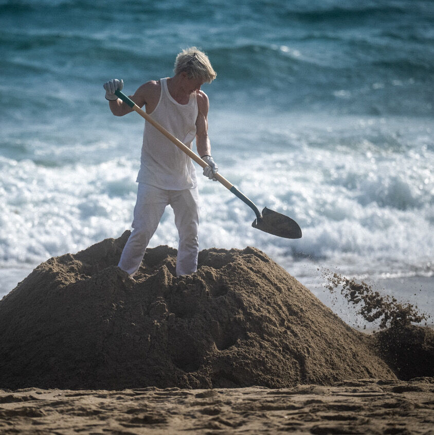 Video still of CASSILS featured work "As It Is". Artist is wearing a white tank top and white pants. They are standing on top of a pile of sand that they are digging out of a beach with the crashing waves of the ocean behind them.