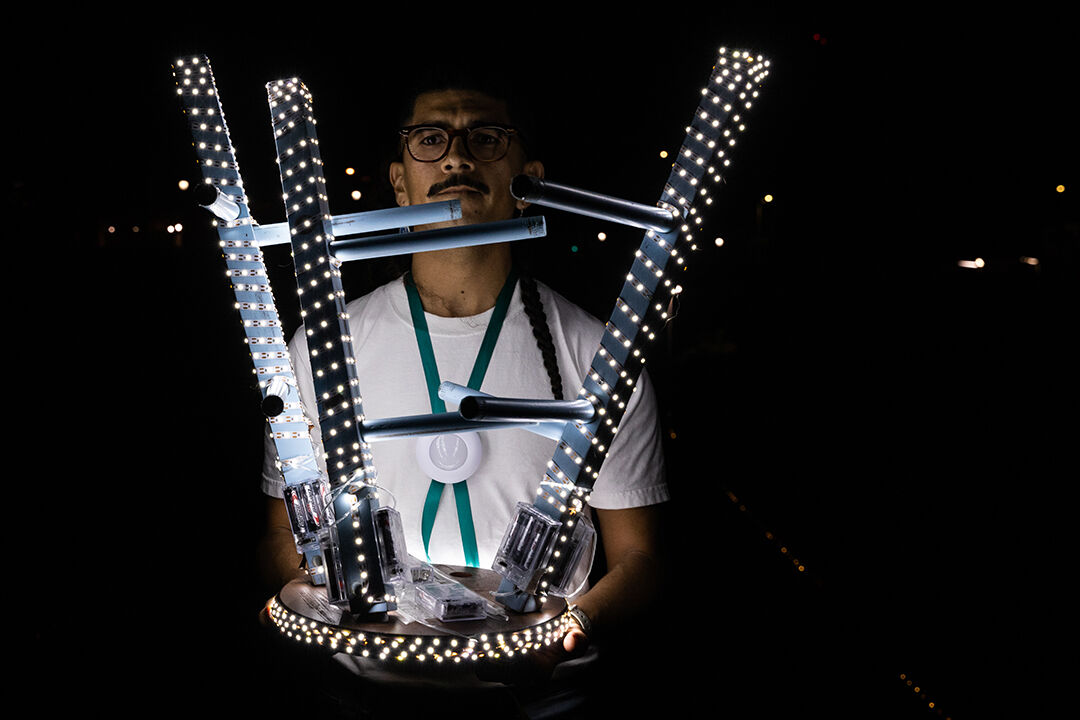 Jose is holding a stool chair that is covered in LED lights wrapped around the legs and the base of the chair. He is surrounded by a dark background.