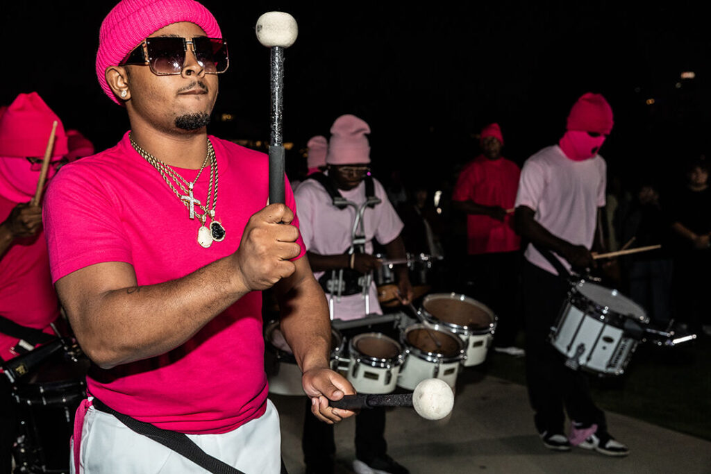 LA Parmelette drum line wearing attire in shades of pink and white while performing at LA Historic Park for XaMENing Masculinities festival. Photo taken at night.