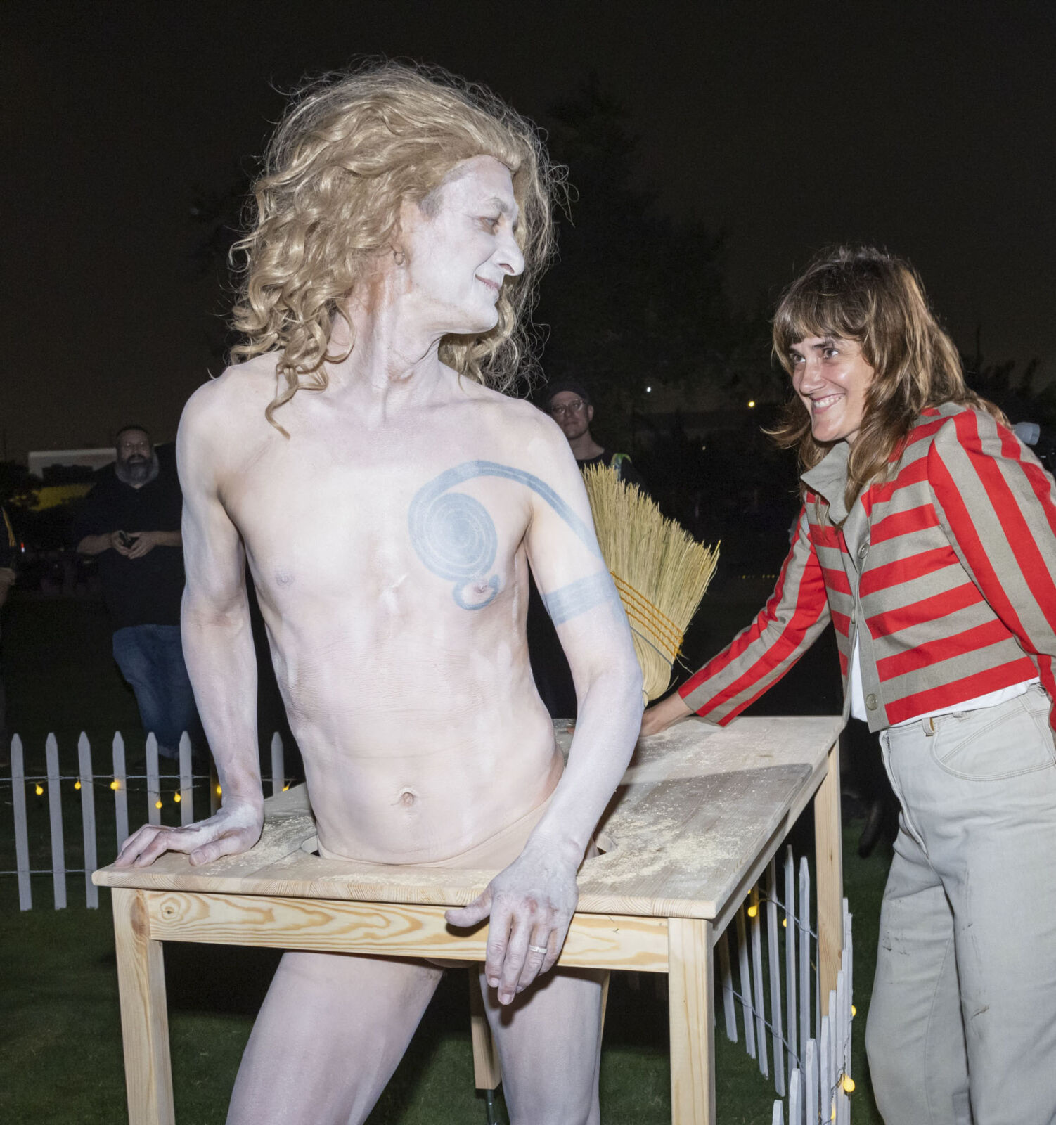 Paul Donald, mostly naked and painted in white with long blonde curly hair, wearing a table around his midsection with a broom attached looking back to an audience member who is touching the table and smiling.
