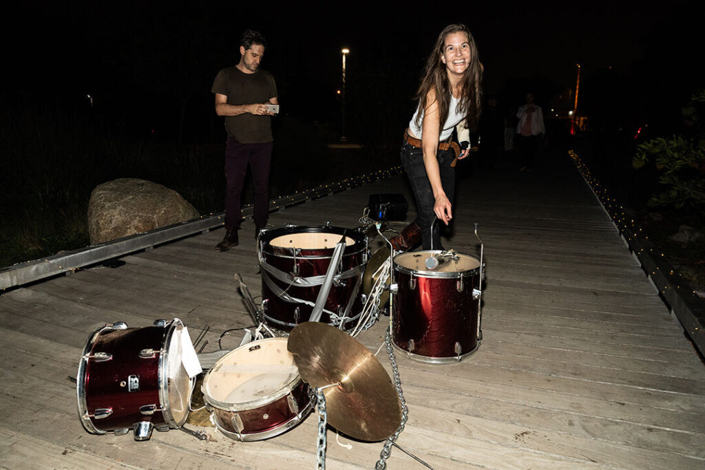Red drumset laying dismantled on a bridge with Sarah Johnson standing over, smiling.