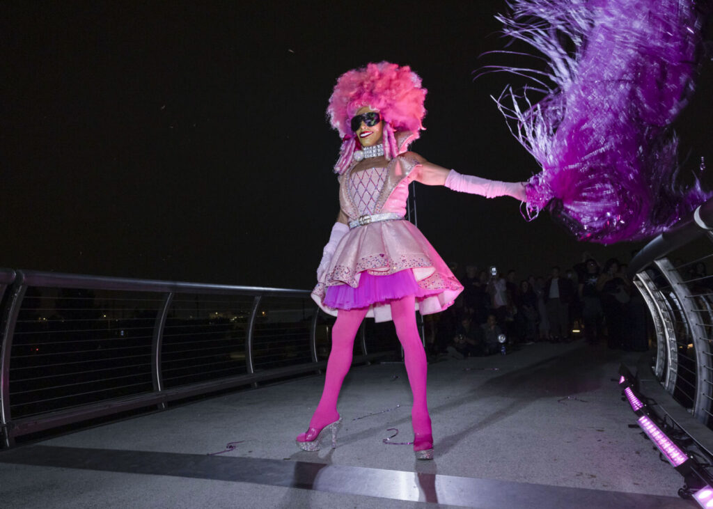 Kiki Xtravaganza wearing entirely pink attire (pink roccocco styled wig, pink dress, neon pink tights, and transparent platform heels). She is standing on the bridge of LA Historic Park and fanning a pink pompom made of reflective pink metallic fringes.