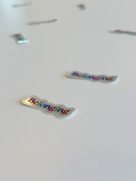 Shinny stickers are scattered on a white table, the stickers has the word "Belonging" - each letter is a different color.