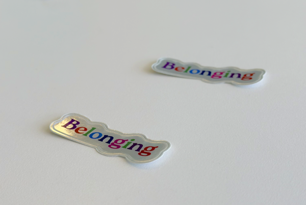 Shinny stickers are scattered on a white table, the stickers has the word "Belonging" - each letter is a different color.