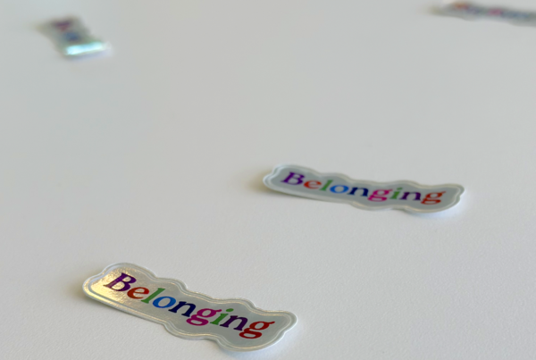 Shinny stickers are scattered on a white table, the stickers has the word "Belonging" - each letter is a different color.